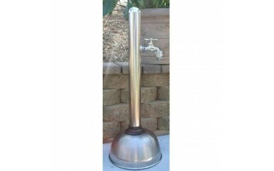 Stainless steel freestanding outdoor shower with curved hose and circular base mounted against stone wall.