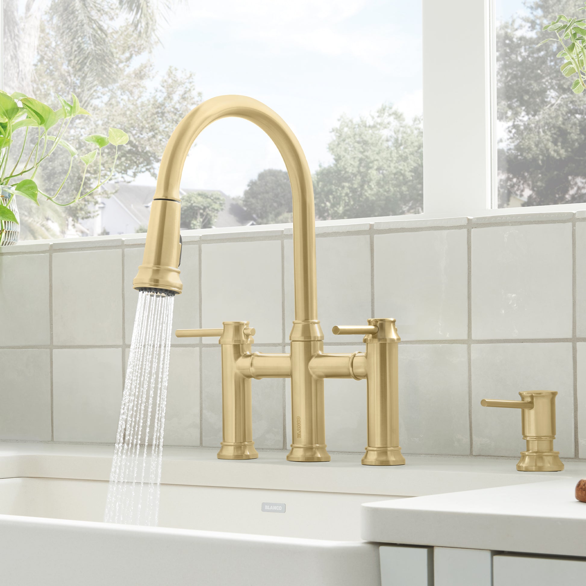 Satin gold pull-down bridge faucet with curved spout, dual handles, and matching soap dispenser on white sink.
