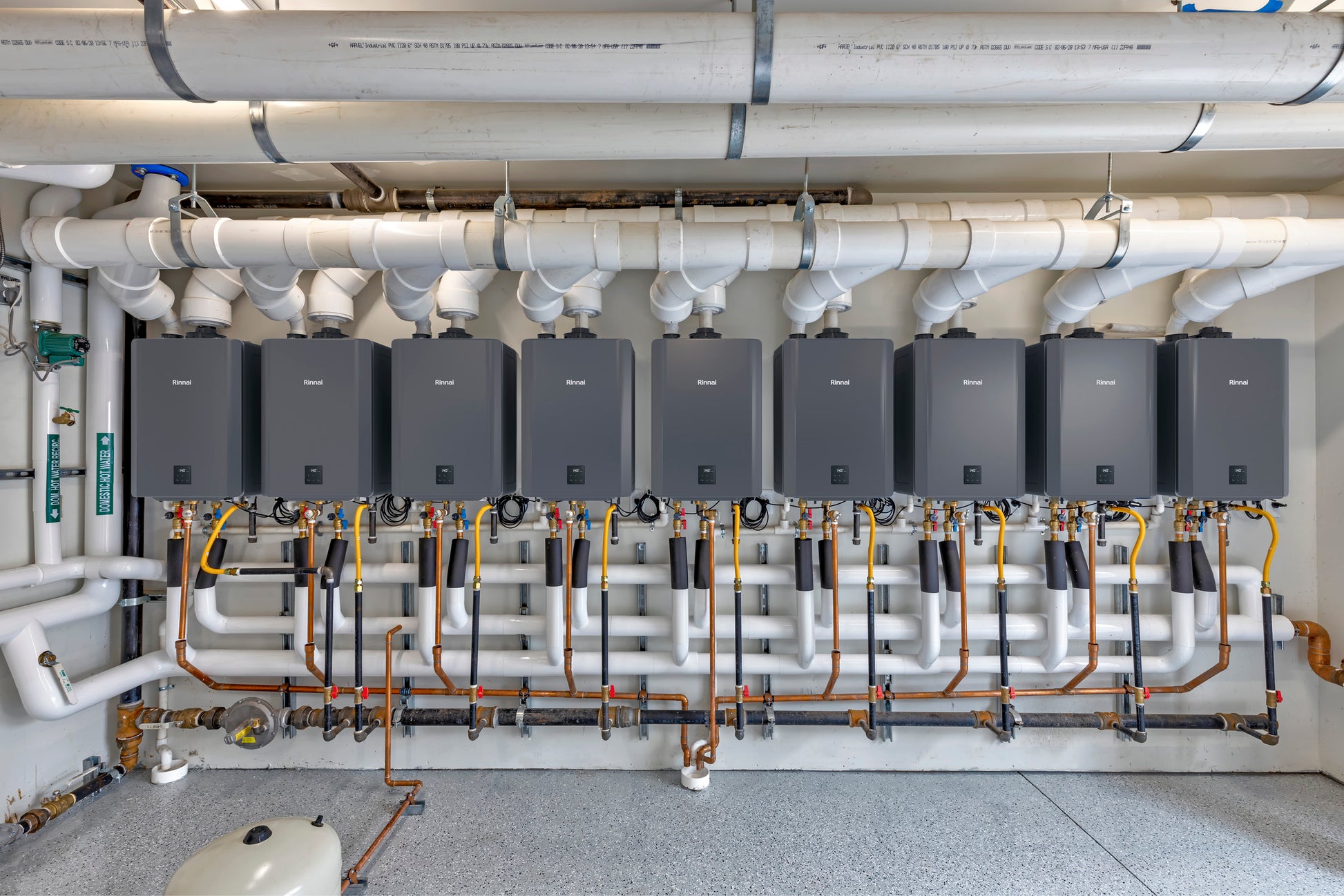 Nine black Rinnai tankless water heaters mounted in a row with white PVC piping and copper connections above.