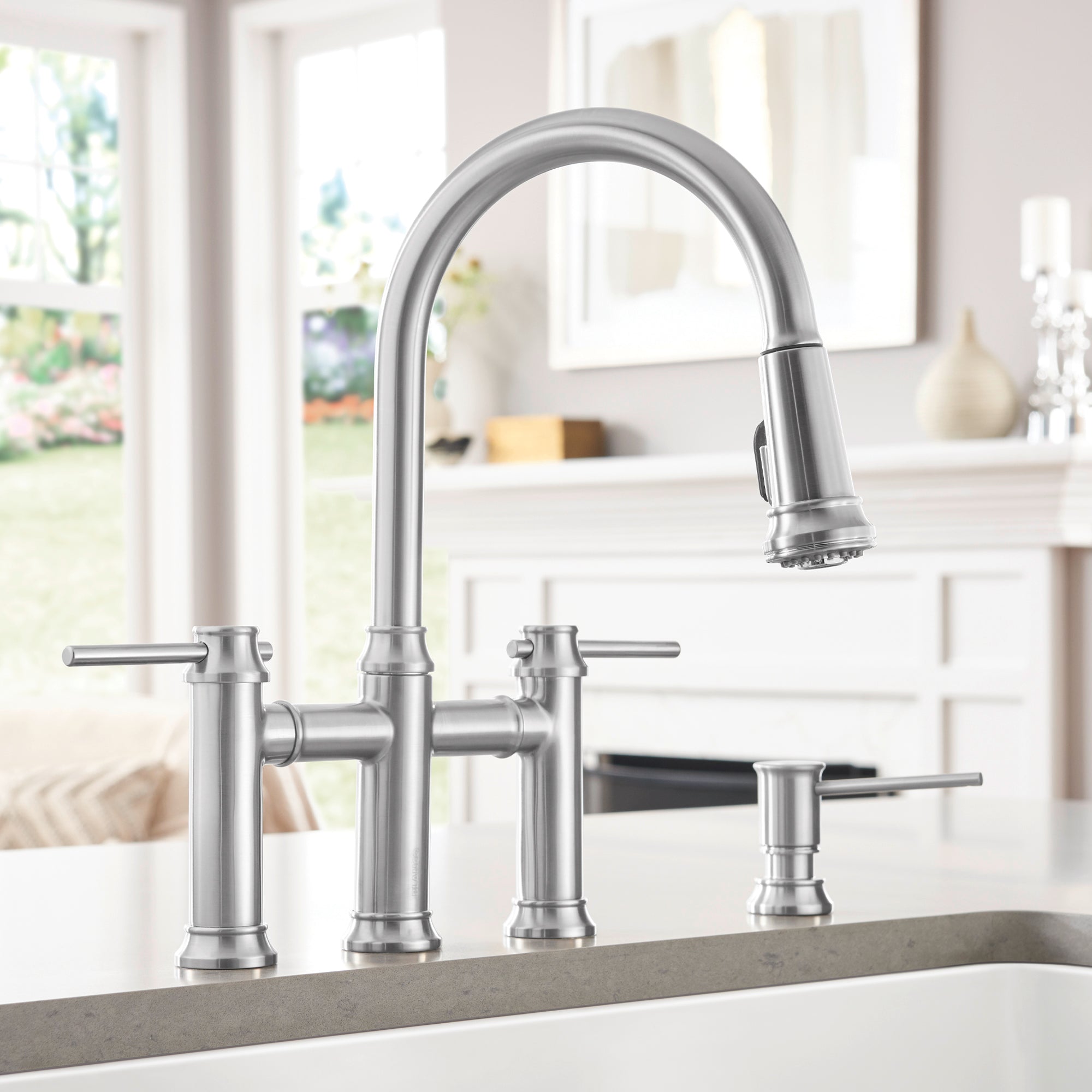 Stainless steel bridge pull-down kitchen faucet with curved spout, dual handles, and separate soap dispenser on countertop.
