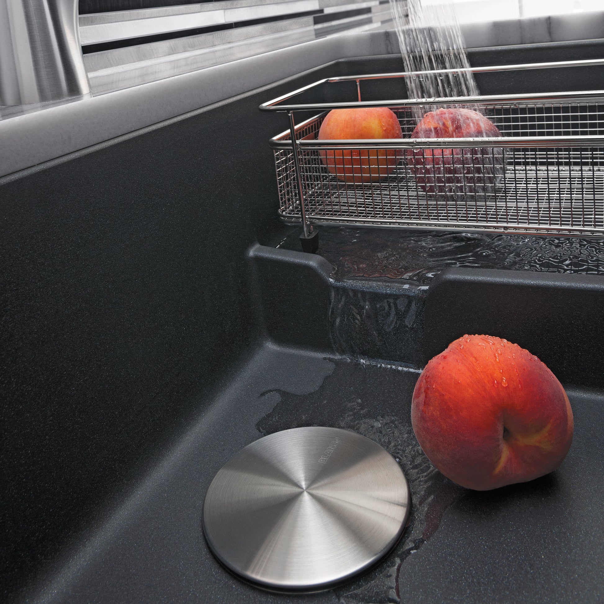 Anthracite undermount sink with cascading water faucet, metal strainer basket, and apples resting in basin.