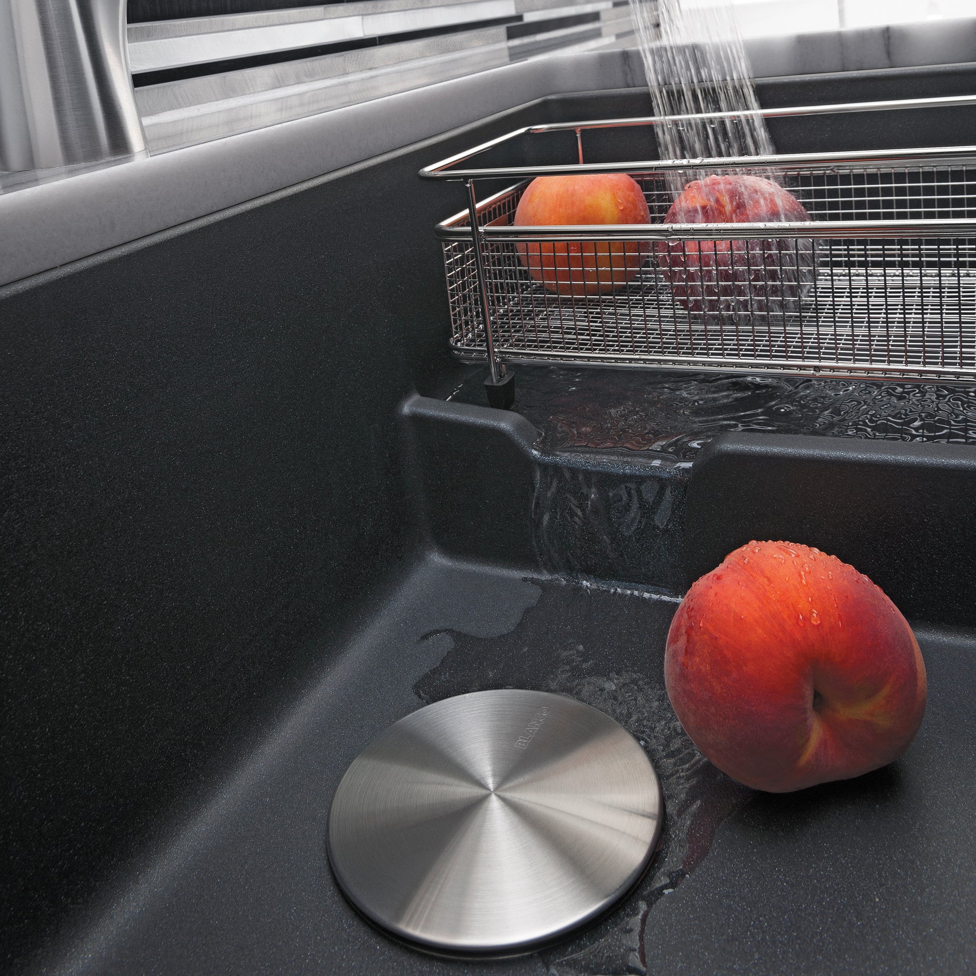 Anthracite undermount sink with cascading water faucet, metal strainer basket, and apples resting in basin.