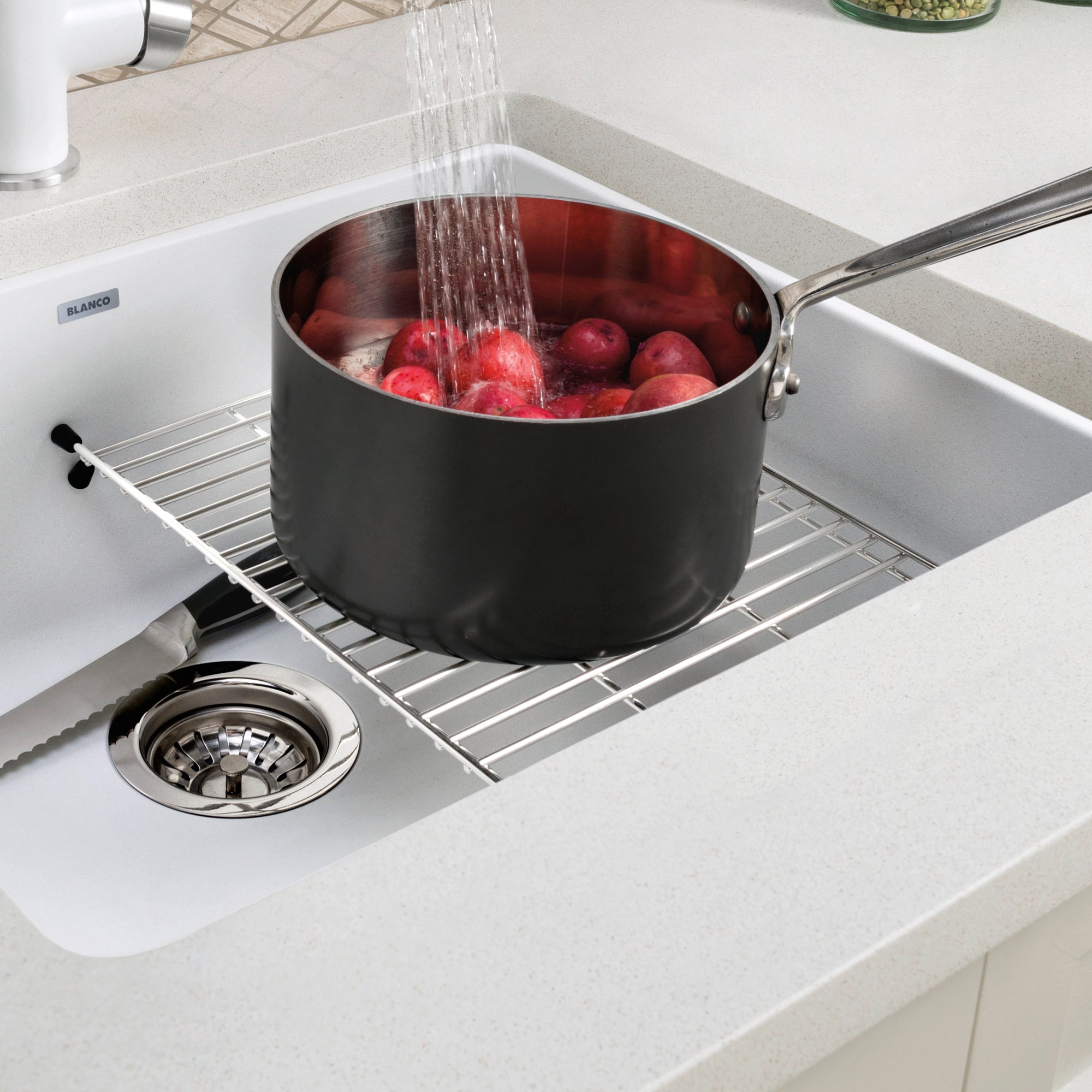 Stainless steel sink grid with parallel bars positioned in white kitchen sink basin with pot of red apples.
