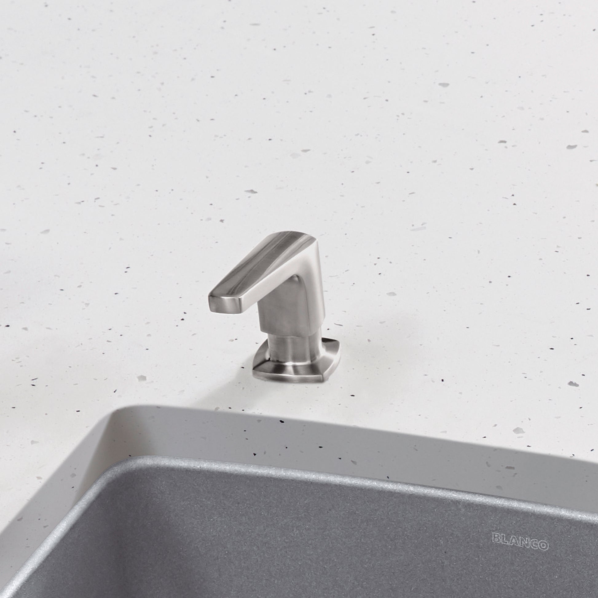 Brushed steel soap dispenser with angled spout mounted on speckled white countertop next to gray sink.
