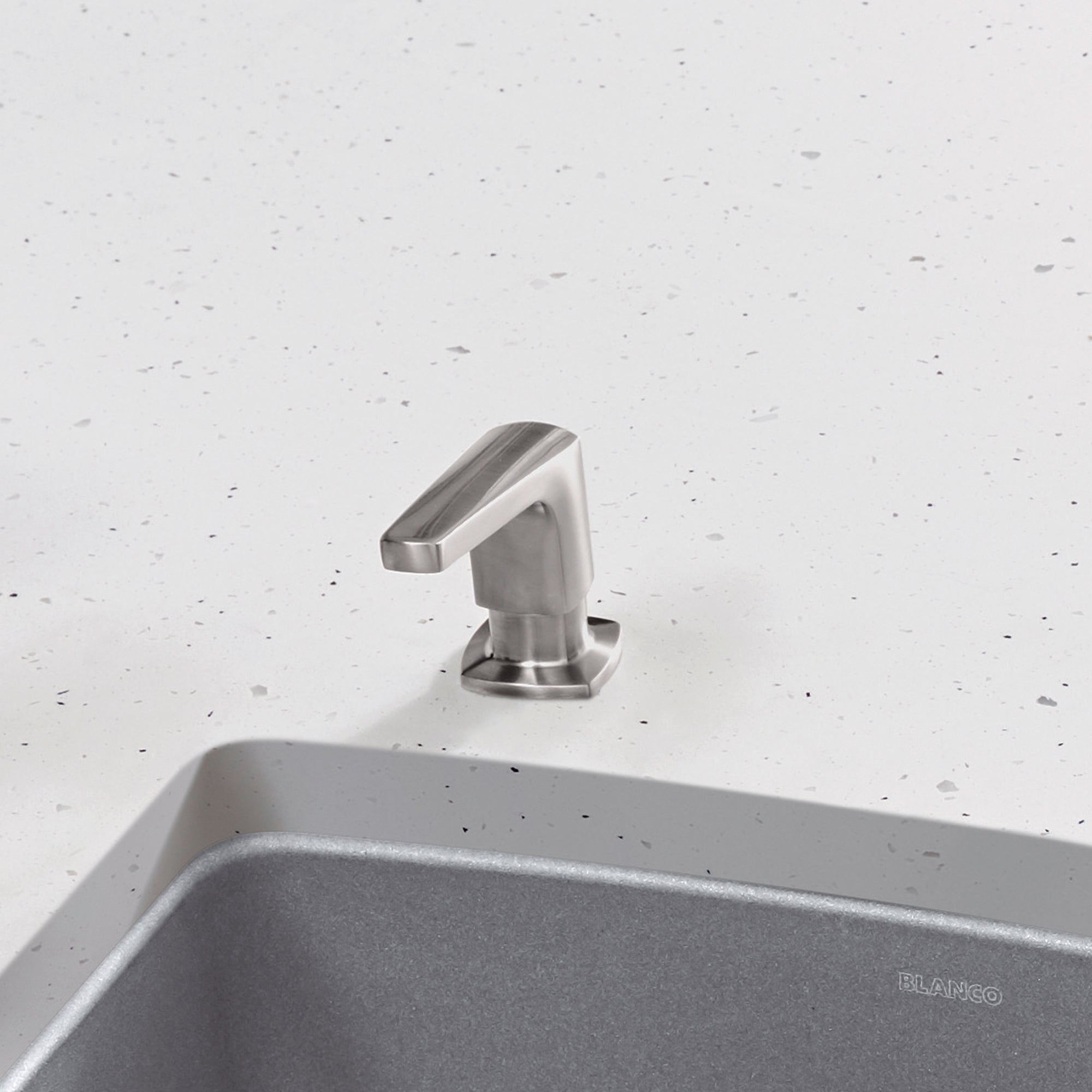 Brushed steel soap dispenser with angled spout mounted on speckled white countertop next to gray sink.