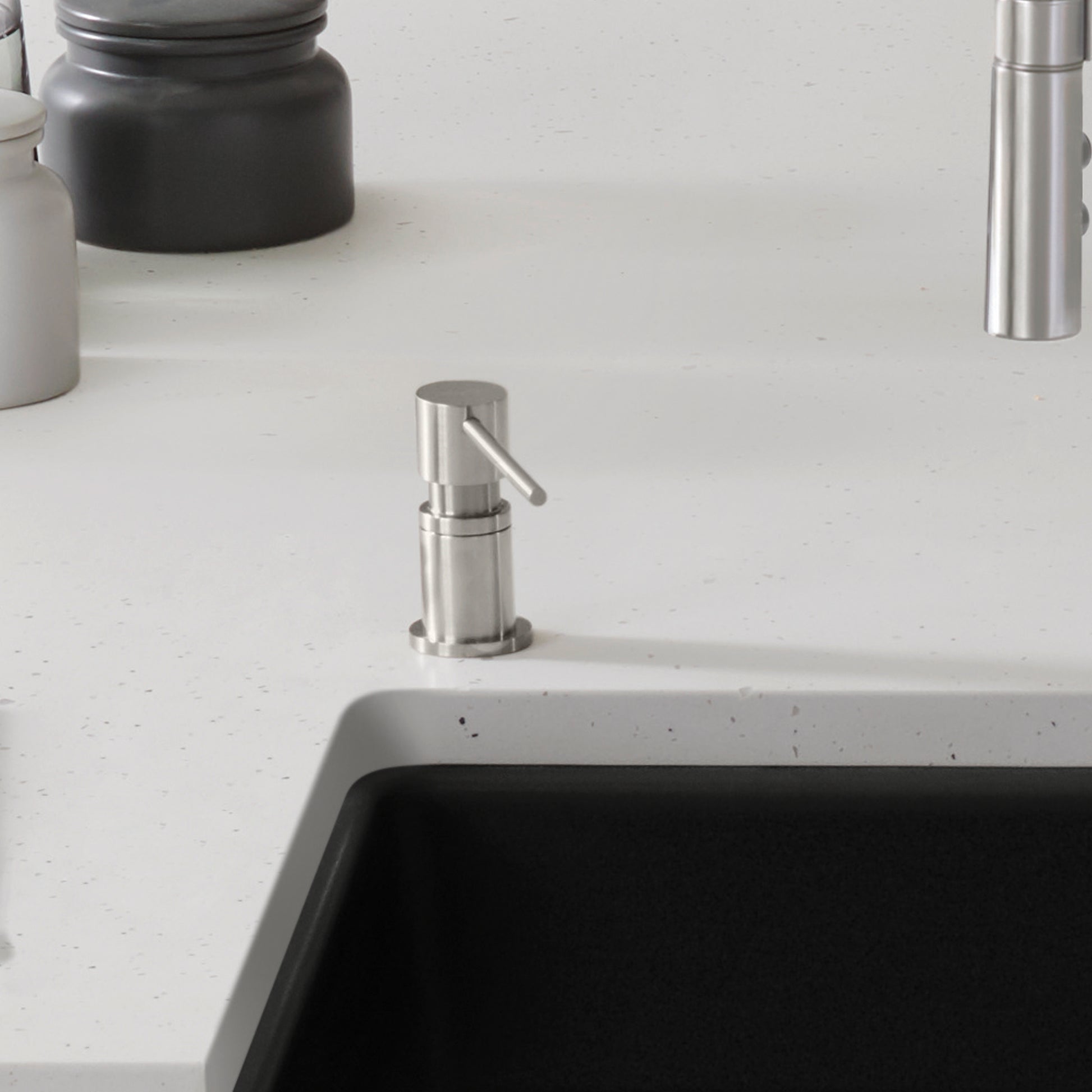 Stainless steel soap dispenser with cylindrical pump and lever, mounted on countertop next to sink.