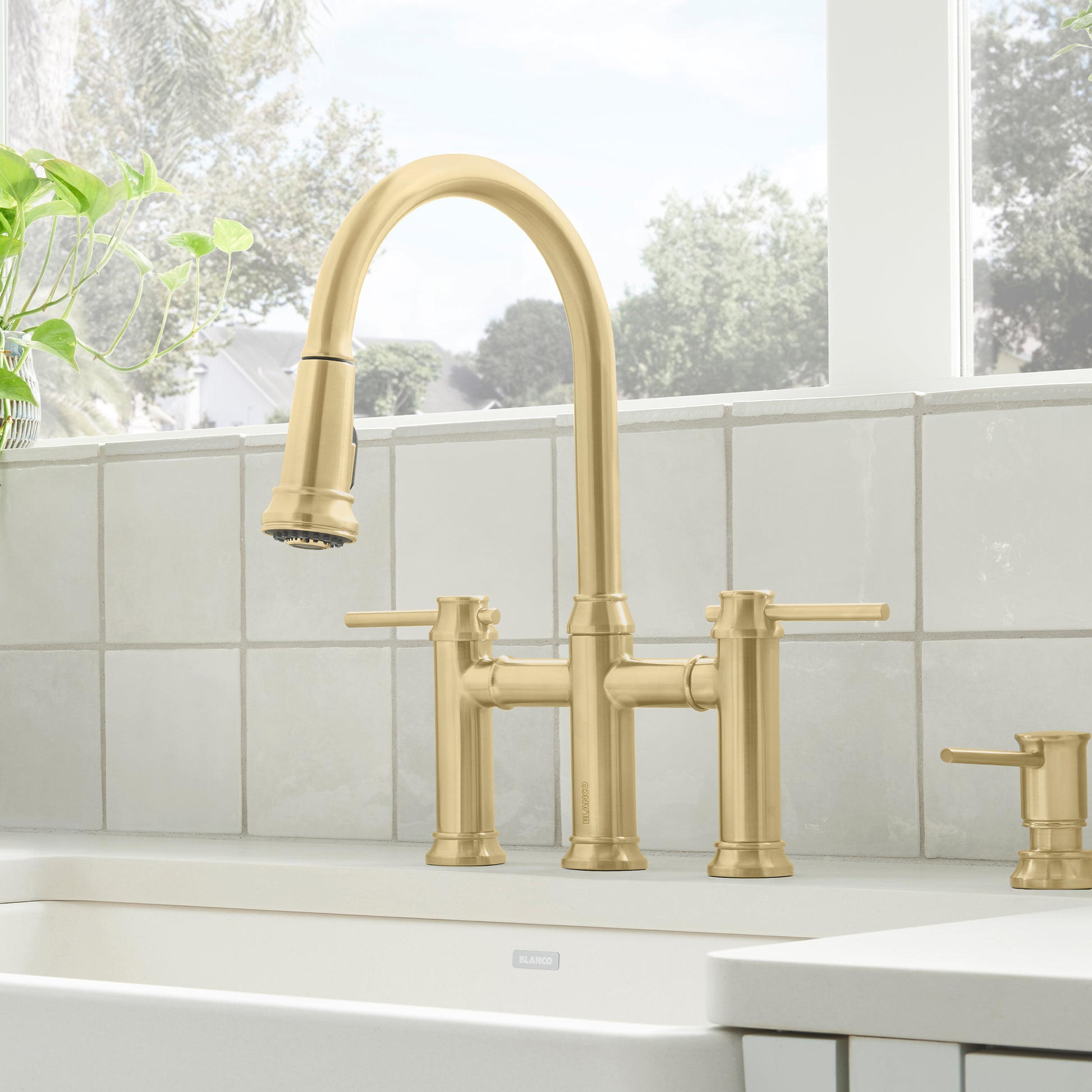 Satin gold bridge faucet with pull-down sprayer, dual lever handles, and matching soap dispenser on white sink.