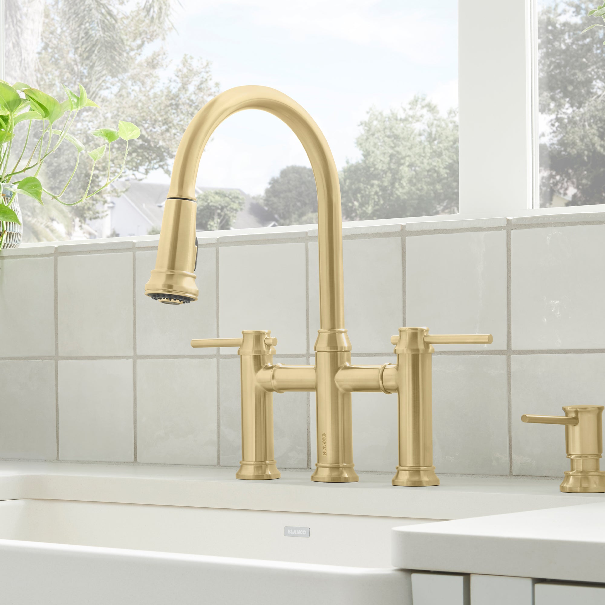 Satin gold bridge faucet with pull-down sprayer, dual lever handles, and matching soap dispenser on white sink.