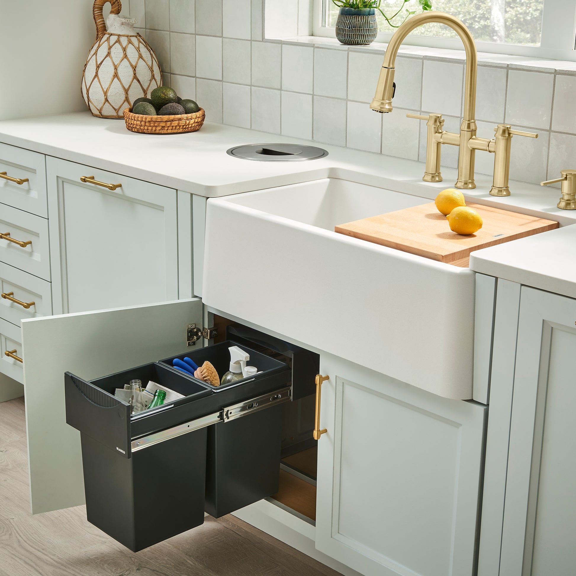 White apron front kitchen sink installed in light green cabinetry with brass faucet and wooden cutting board.