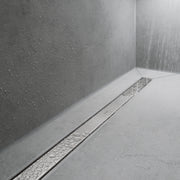Brushed stainless steel linear floor drain installed in white tile shower with water flowing down textured gray walls.