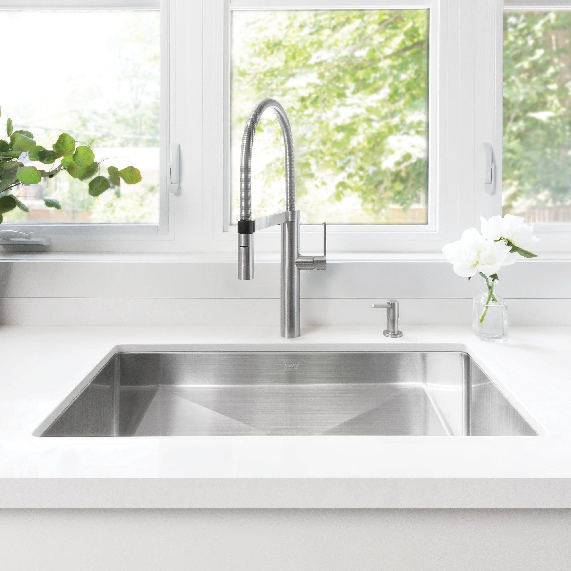 Stainless steel single bowl undermount sink installed in white countertop with arched faucet and soap dispenser.