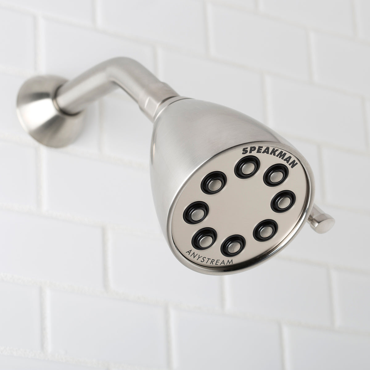 Brushed nickel shower head with round face featuring eight spray holes, mounted on angled arm against white tile wall.