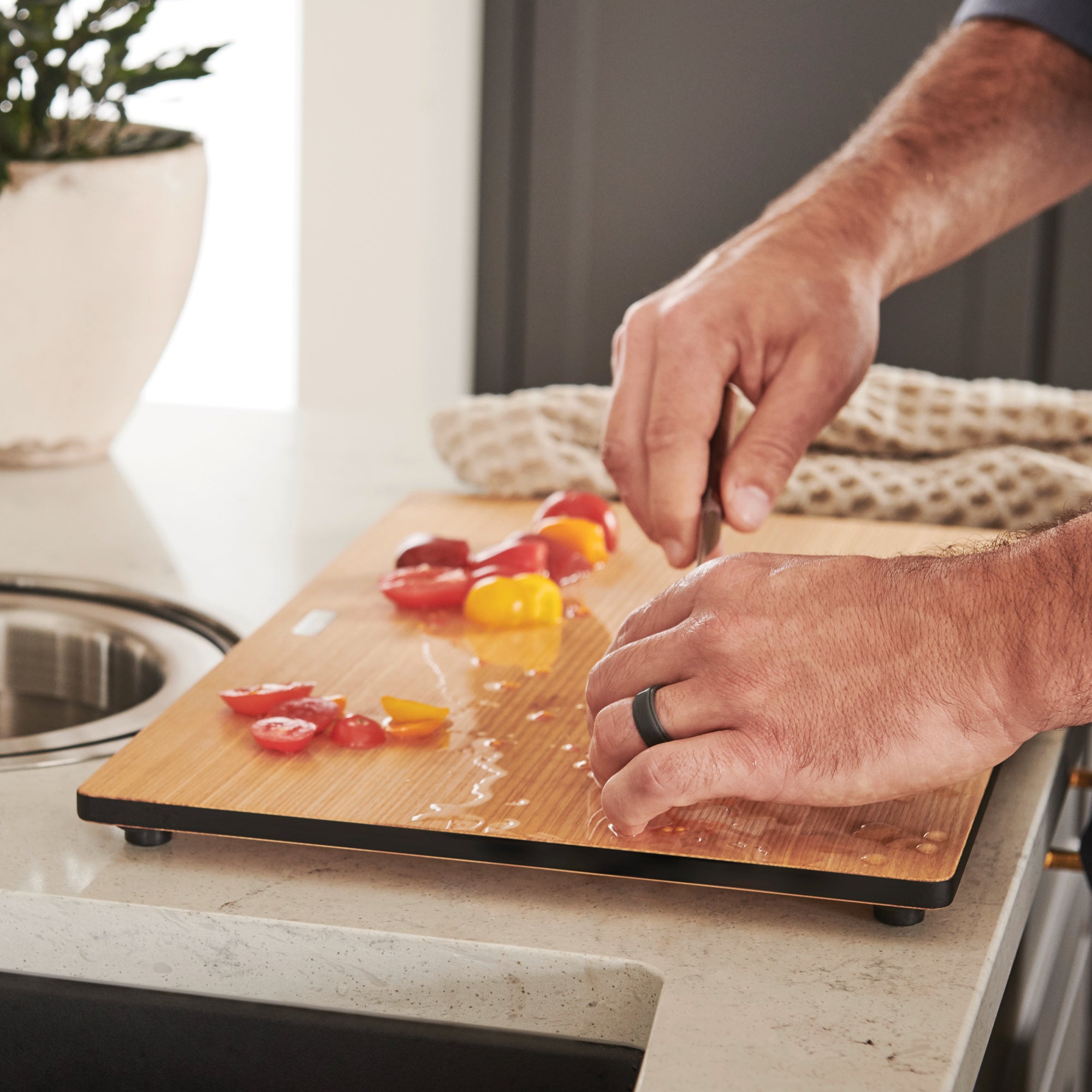Light wood cutting board with black edge and rubber feet, person chopping red and yellow peppers on countertop.