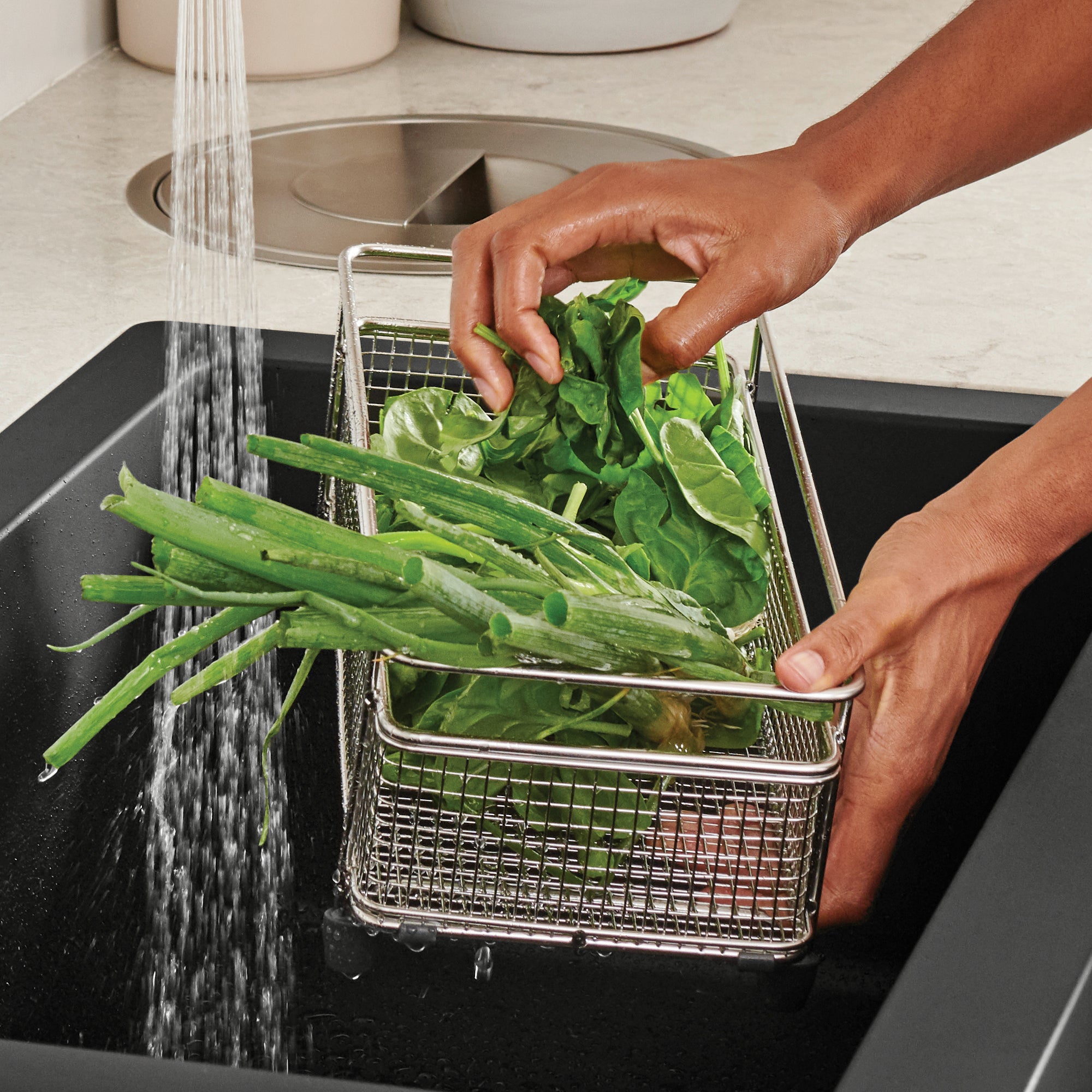 Stainless steel mesh basket holding green beans and leafy greens under running water in a sink.