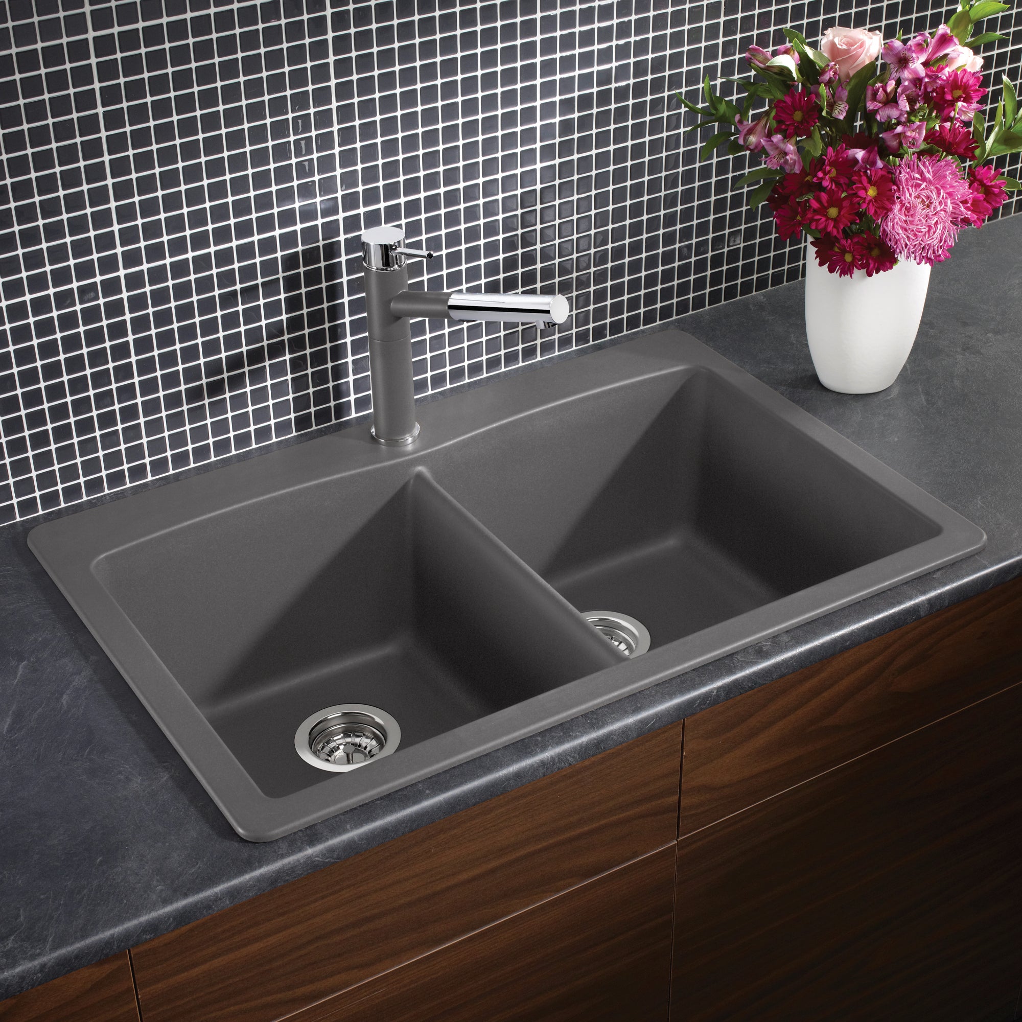 Cinder gray double bowl kitchen sink with chrome faucet installed in dark countertop and wood cabinetry.