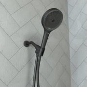 Matte black handheld showerhead with circular perforated face mounted on adjustable holder against herringbone tile wall.