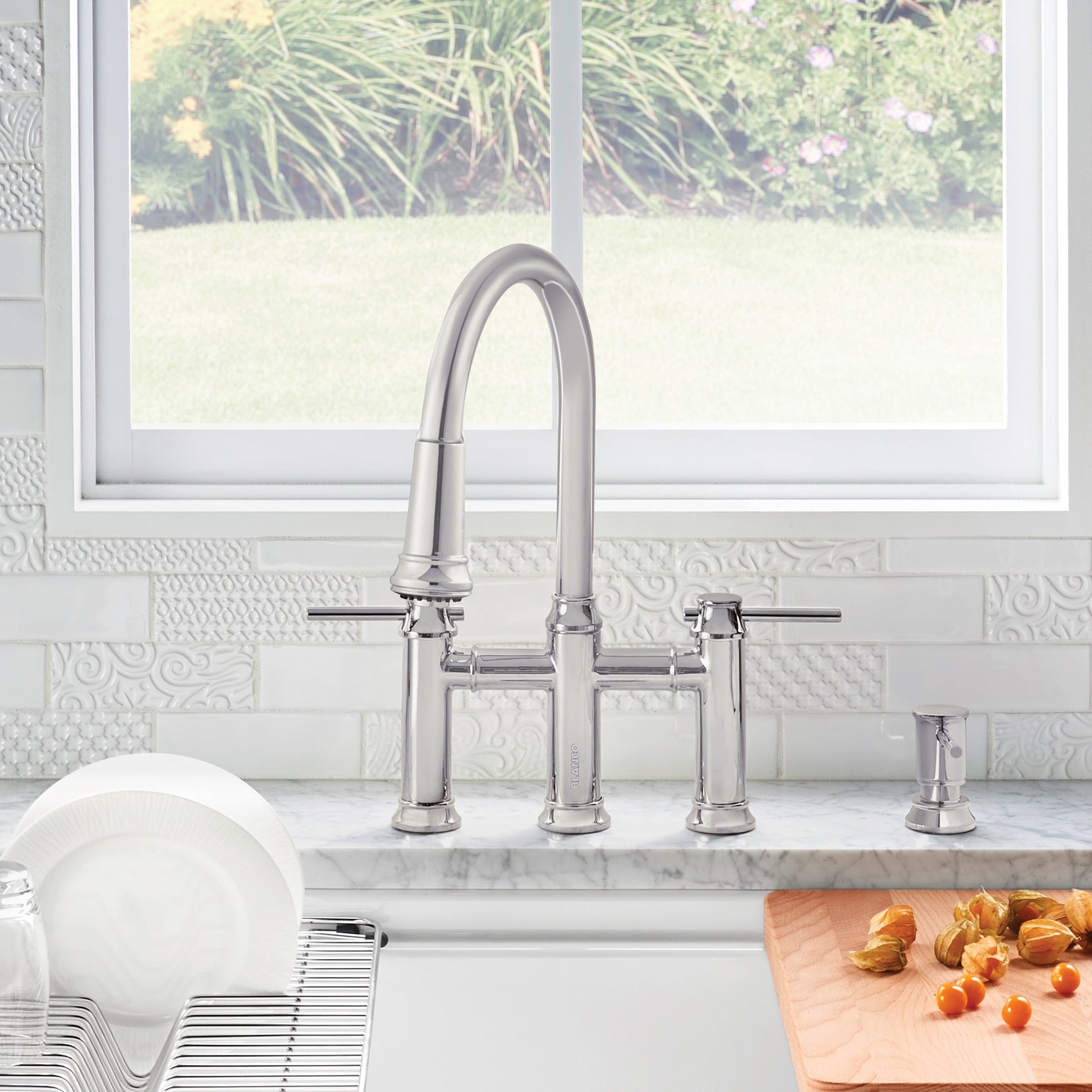 Chrome bridge kitchen faucet with curved pull-down spout, dual handles, and matching soap dispenser on marble countertop.