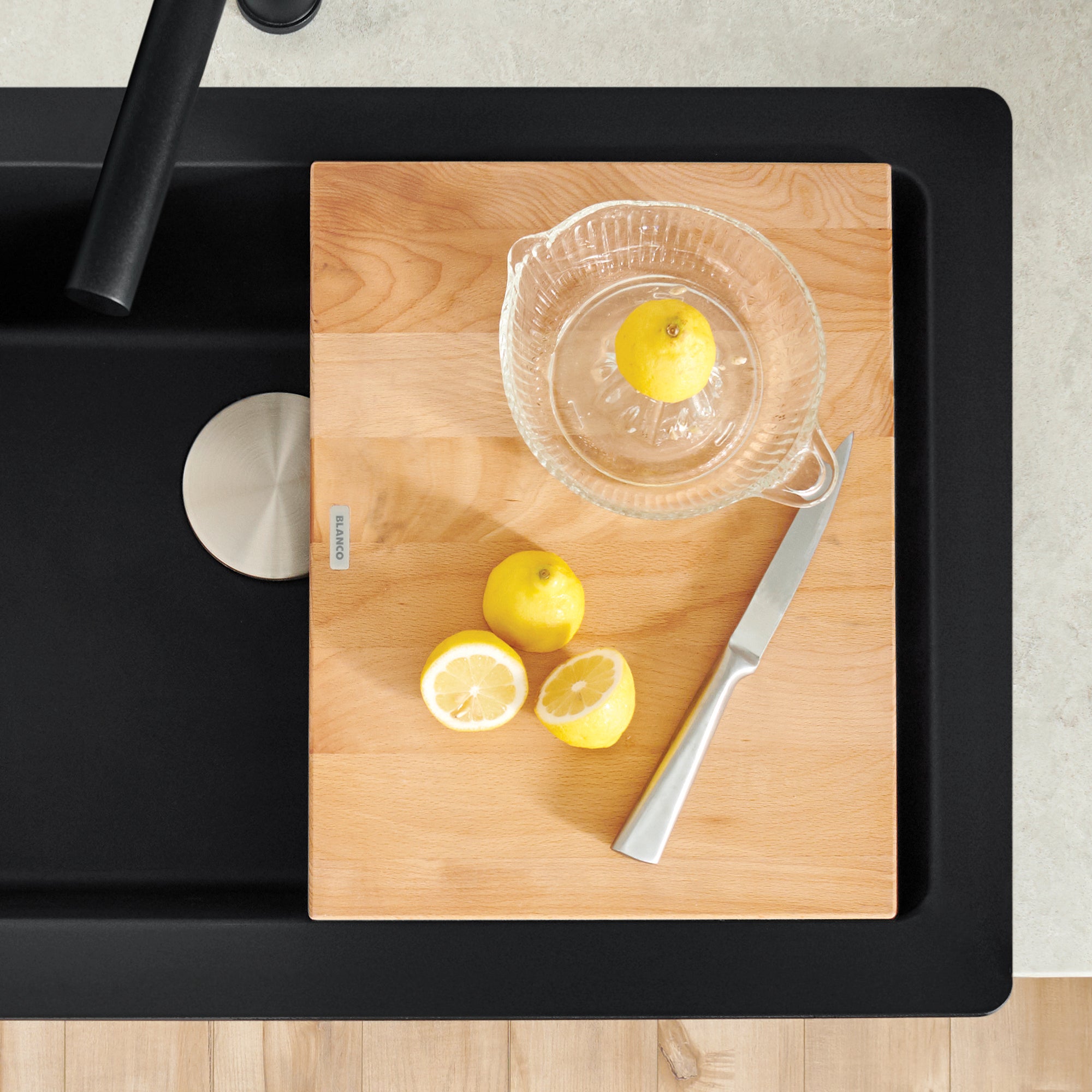 Beechwood cutting board with light wood grain sitting on black sink rim, with lemons and juicer on surface.