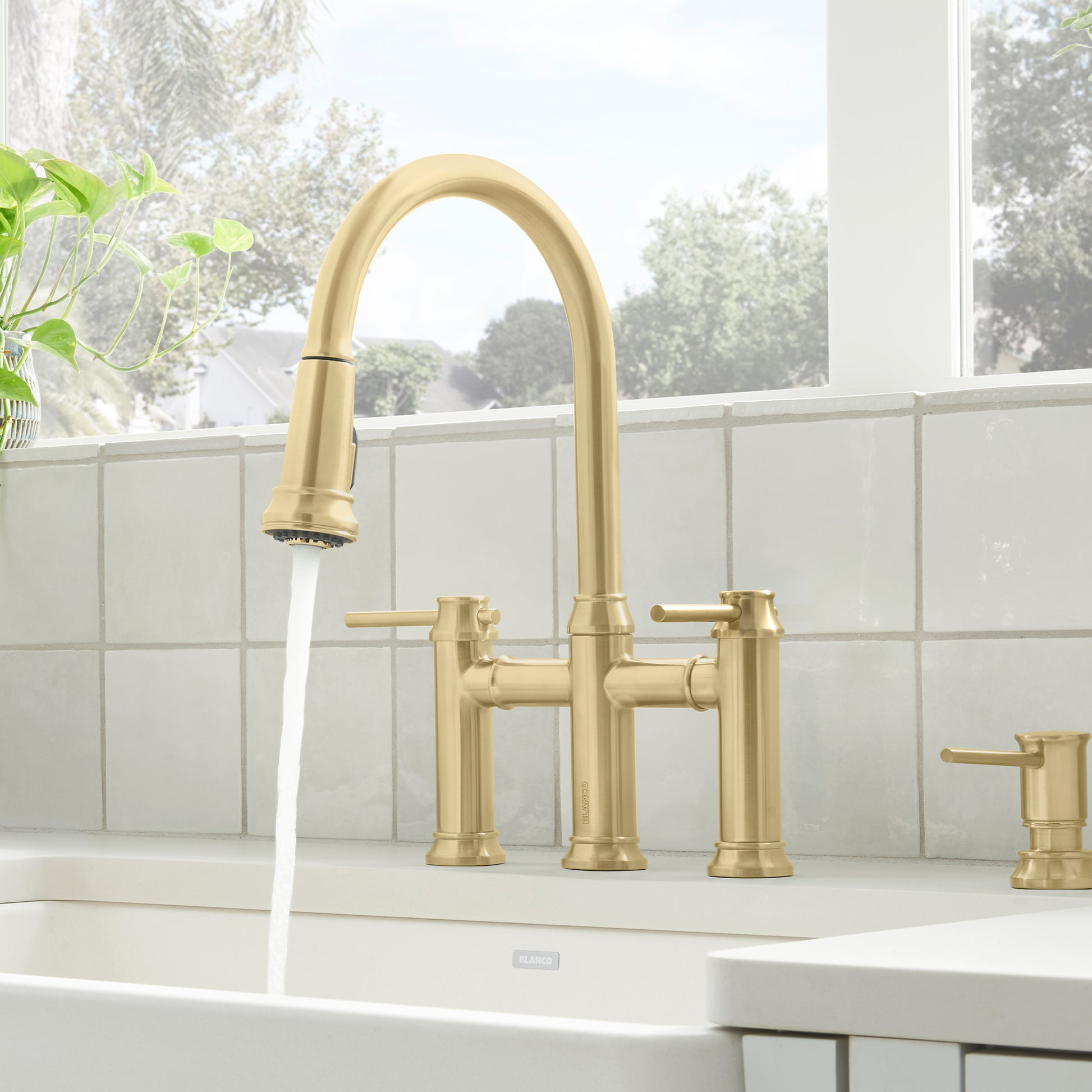 Satin gold pull-down bridge faucet with dual handles and arched spout installed on white sink countertop.