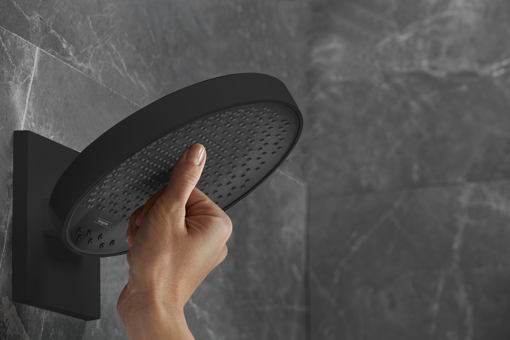 Matte black round shower head with perforated face mounted on wall with hand touching surface.
