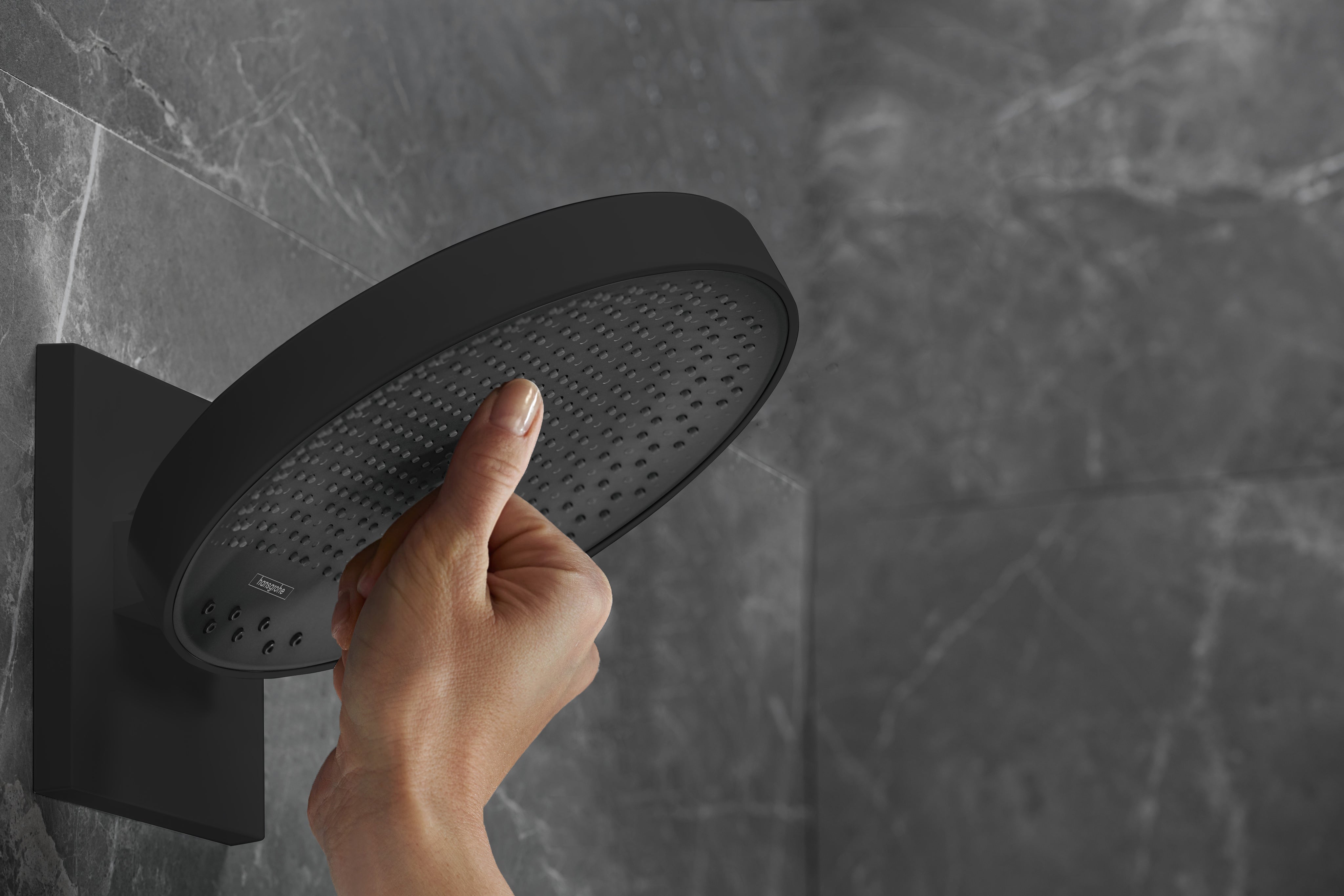 Matte black round shower head with perforated face mounted on wall with hand touching surface.