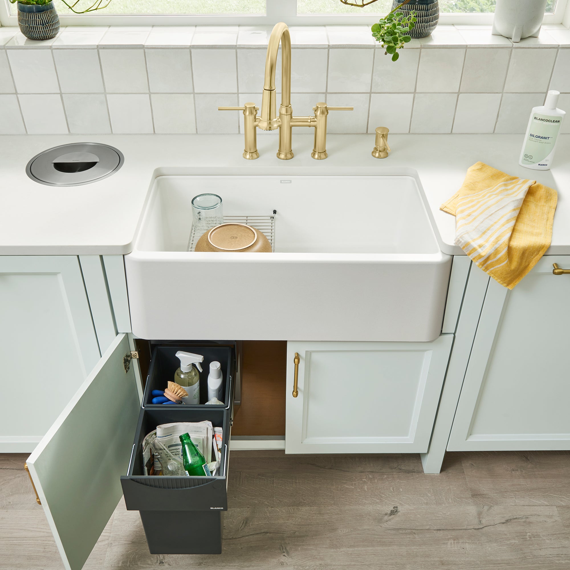 White farmhouse sink with brass faucet, white countertop, and black pull-out storage system containing cleaning supplies and 