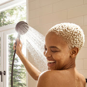 Matte black handheld showerhead with hose held up at an angle, dispensing water spray in a bathroom with tile walls.