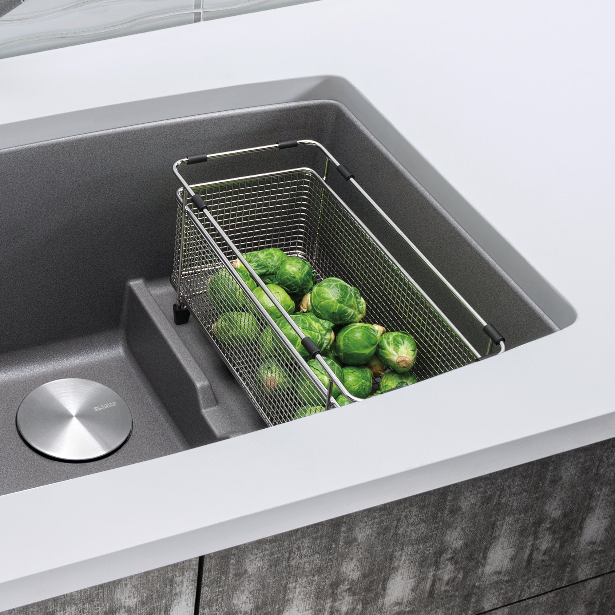 Stainless steel mesh colander with handles sitting in dark gray kitchen sink filled with brussels sprouts.