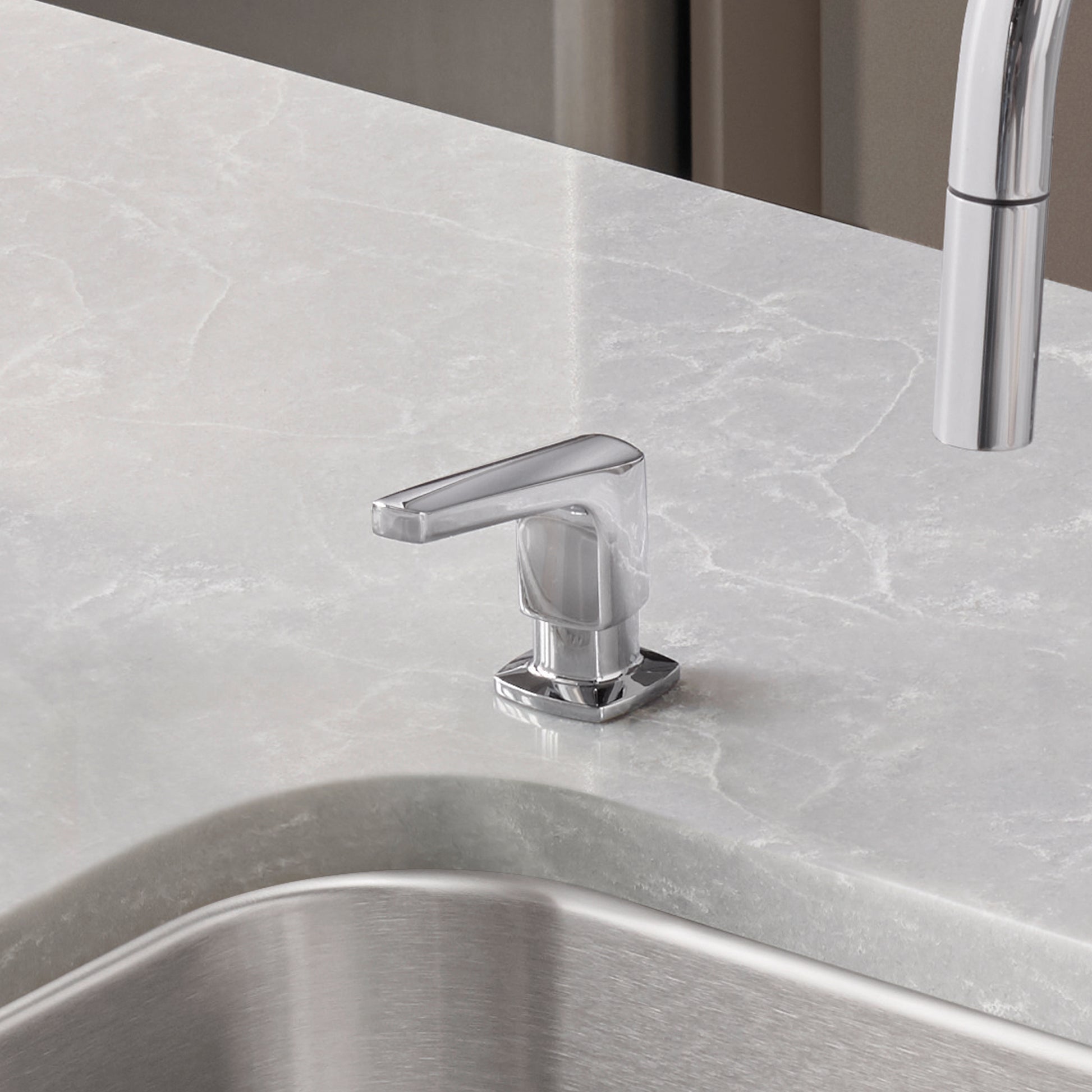 Chrome soap dispenser with angular spout mounted on marble countertop next to stainless steel sink.