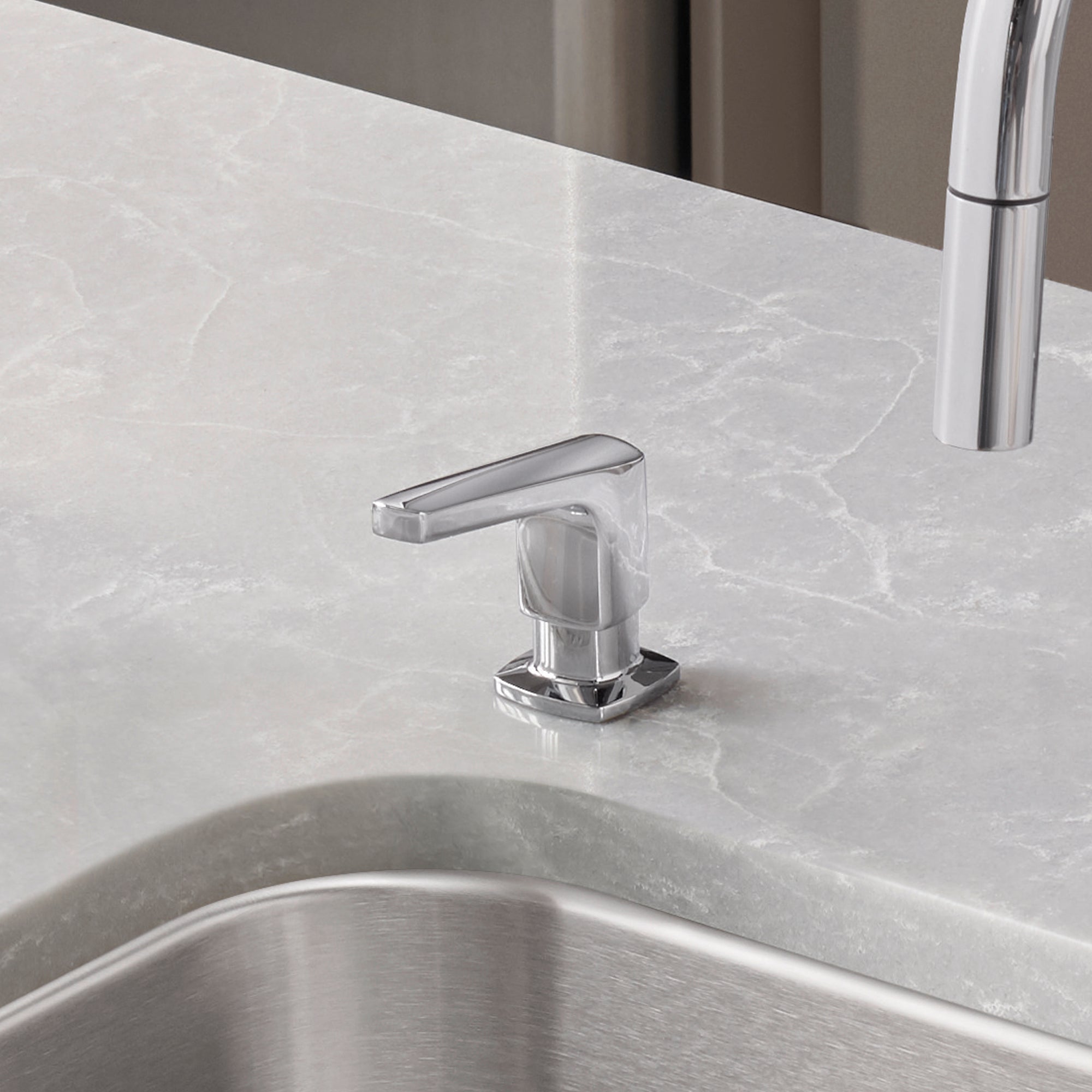 Chrome soap dispenser with angular spout mounted on marble countertop next to stainless steel sink.