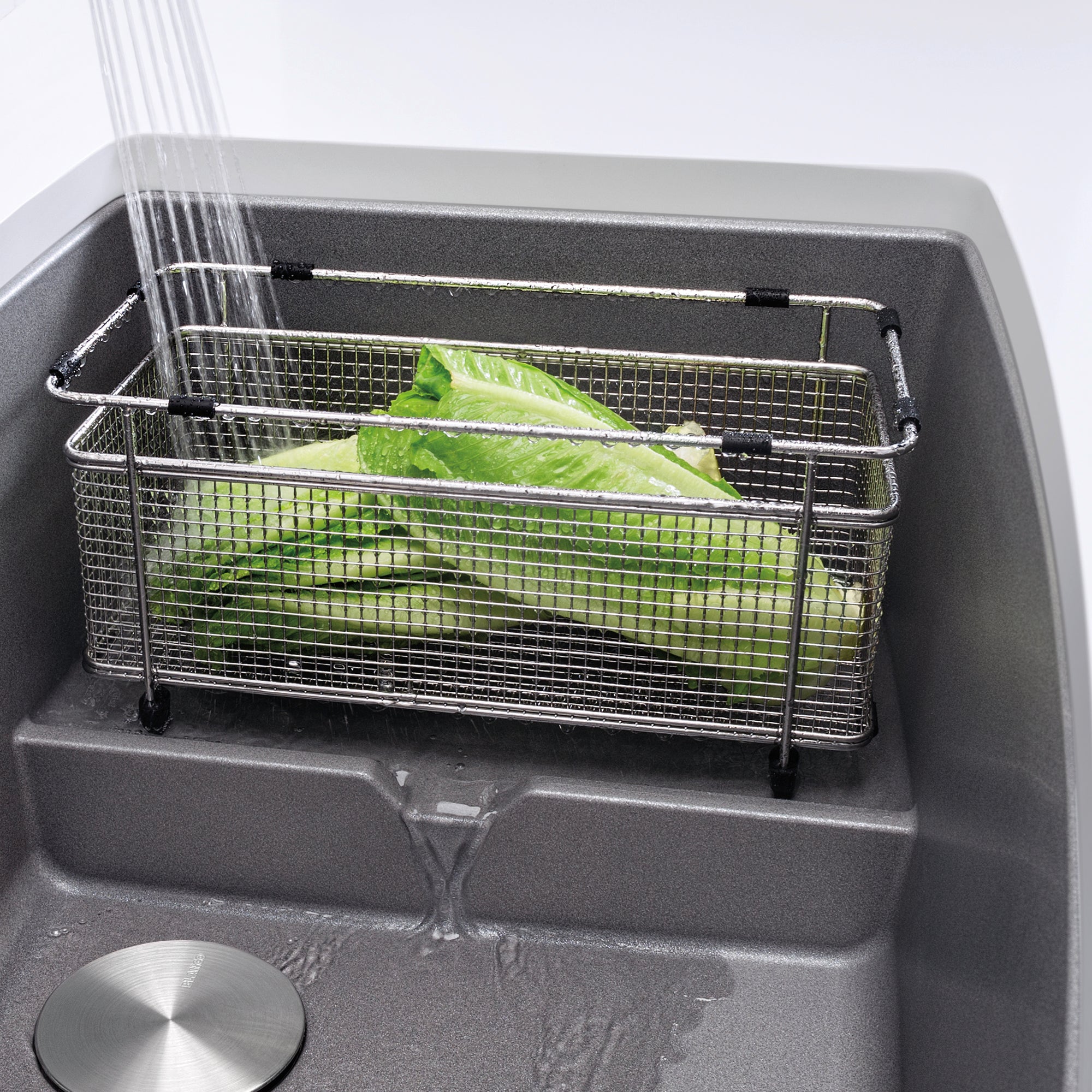 Stainless steel mesh colander with handles positioned in gray sink basin, containing fresh lettuce with water draining.