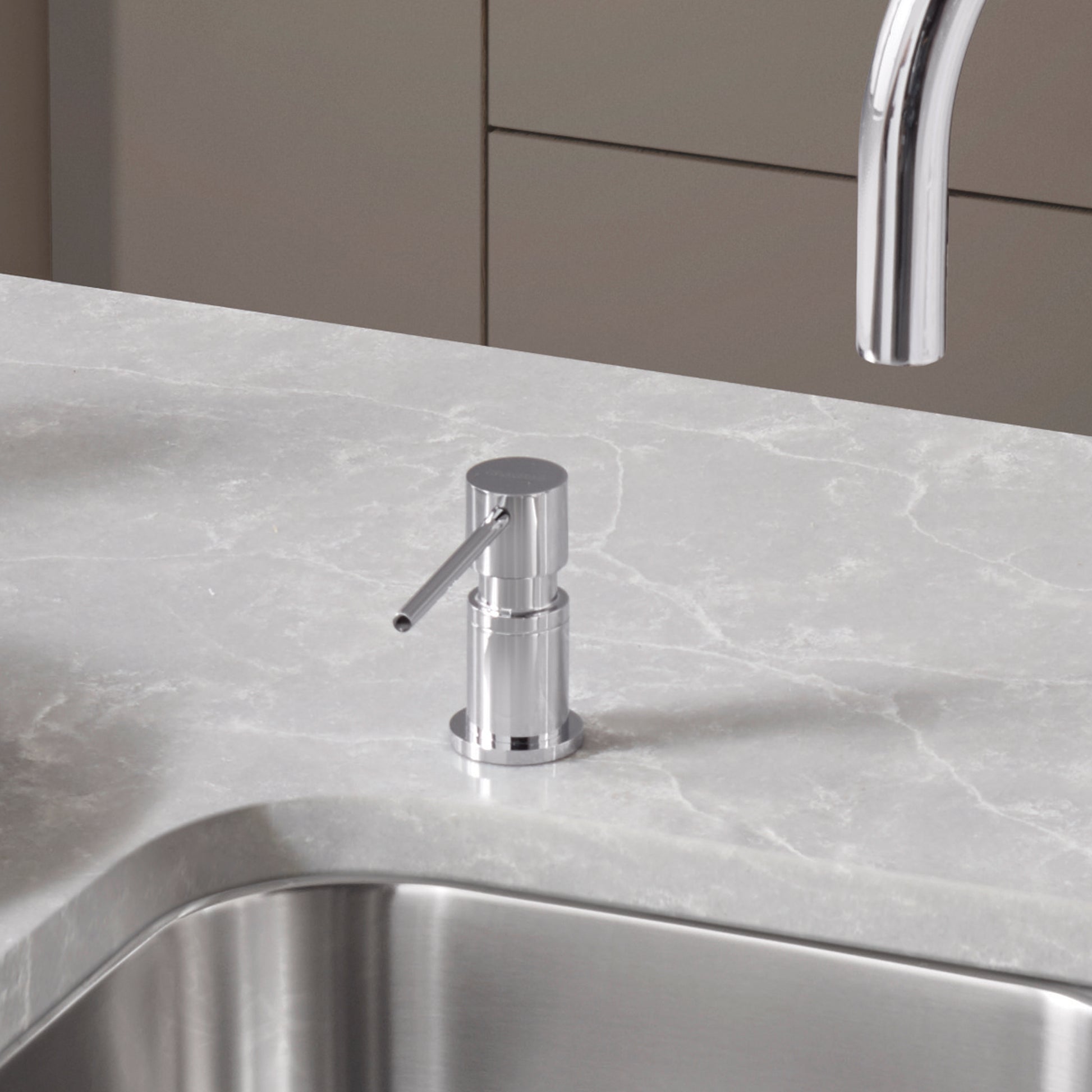 Chrome soap dispenser with cylindrical body and angled spout mounted on marble countertop next to sink.