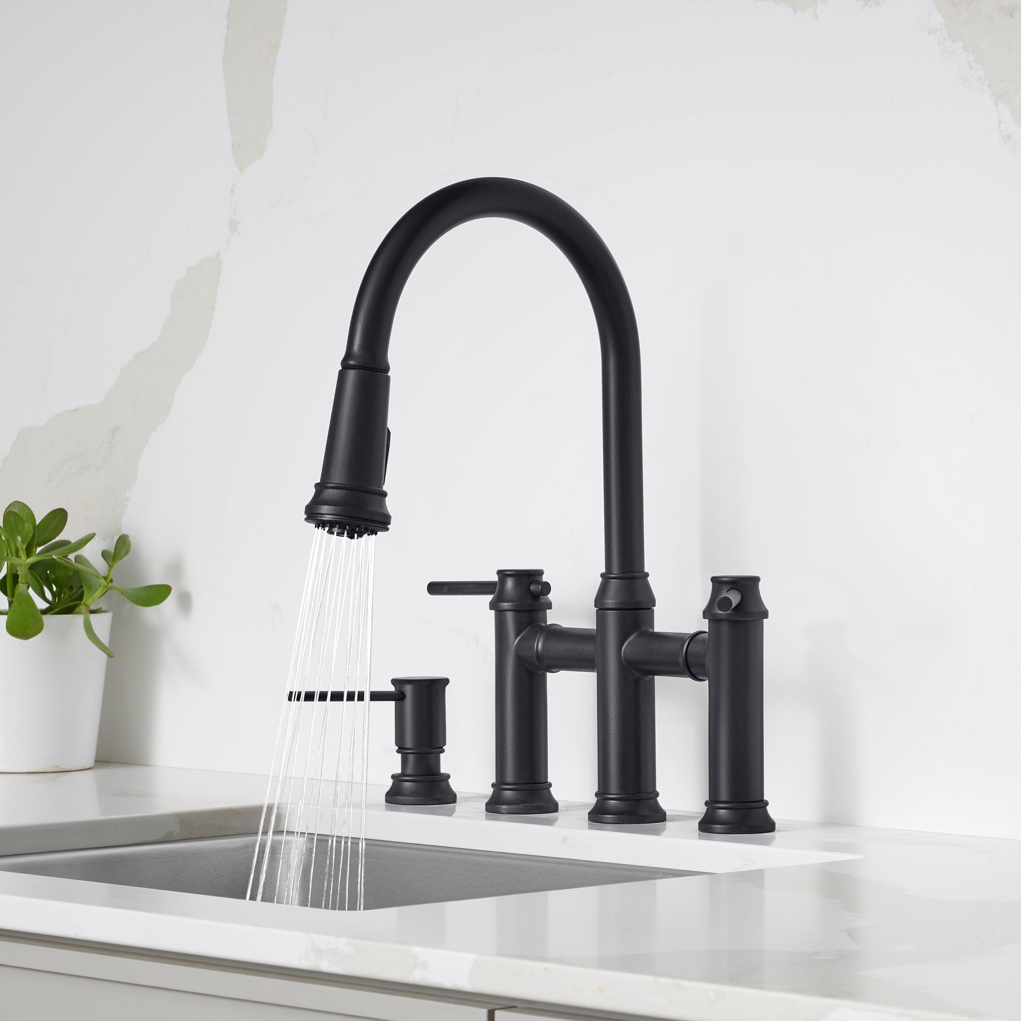 Matte black bridge faucet with pull-down sprayer head, dual handles, and soap dispenser mounted on white countertop sink.
