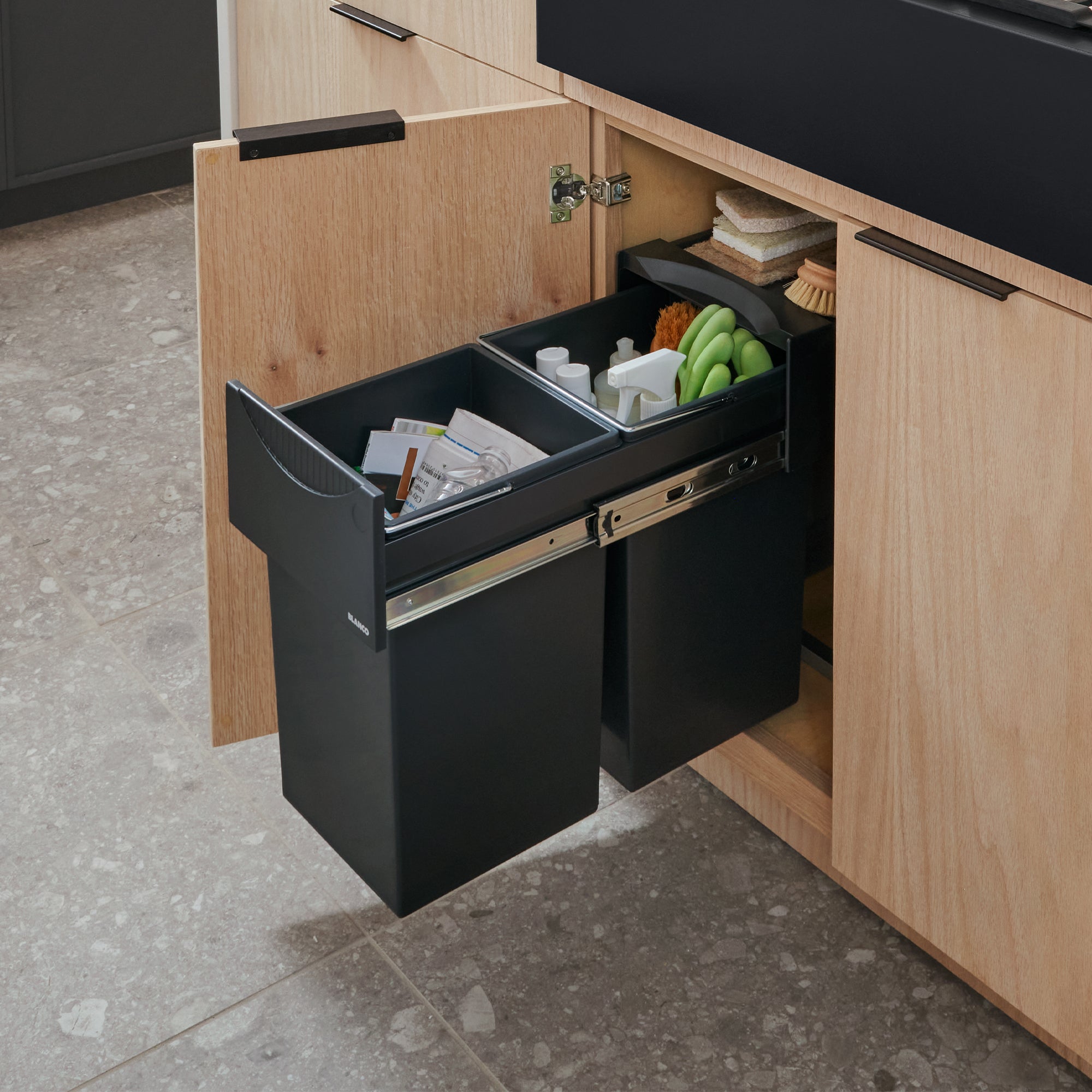Black pull-out waste and recycling storage system with dividers, installed in light wood cabinetry.