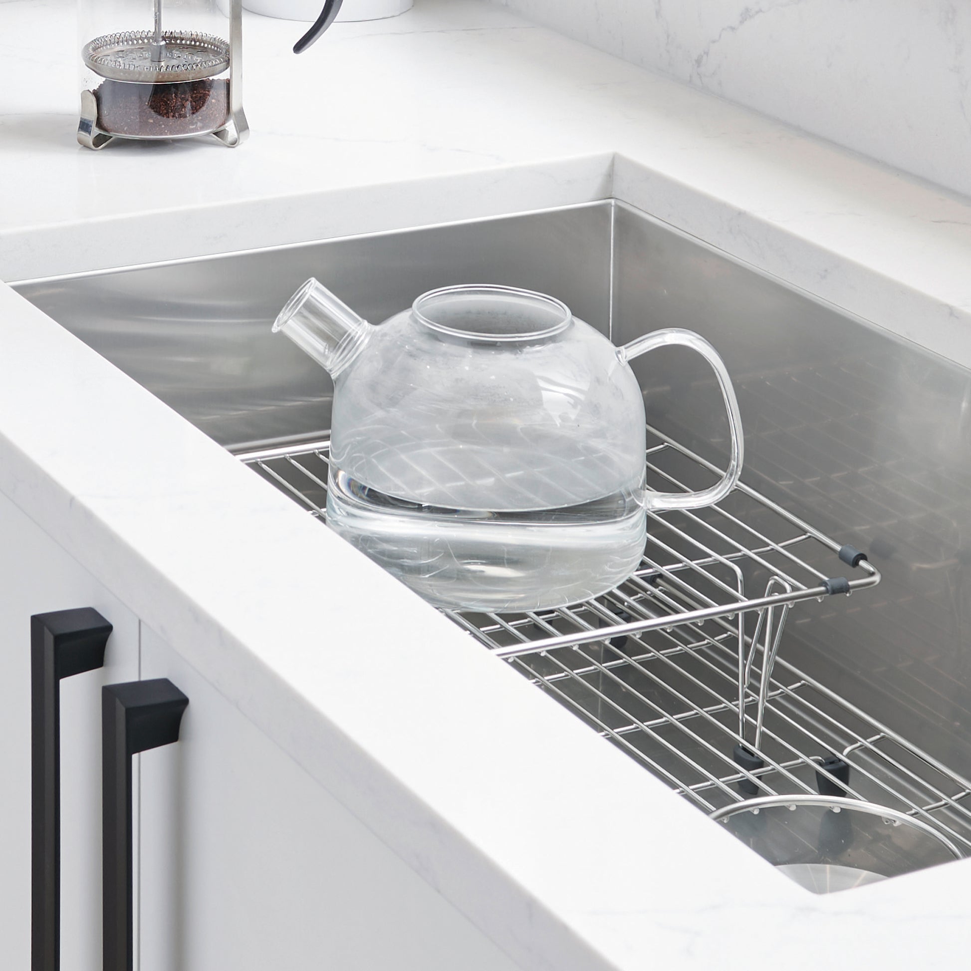 Stainless steel multi-level sink grid with tiered shelves holding a glass teapot, installed in a single bowl sink.