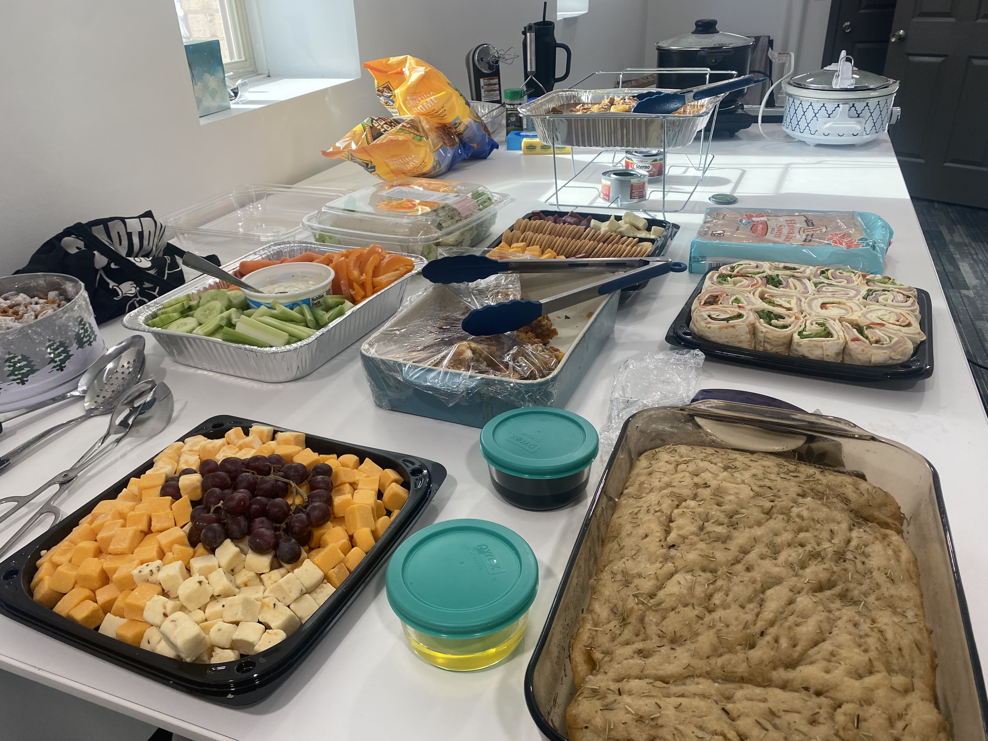 A table spread with homemade focaccia bread, sausage balls, spinach artichoke dip, Mississippi pork roast, mashed potatoes, Hawaiian rolls, cheese, fruit, and salad